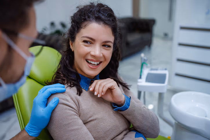 A female patient smiling in a dental chair while a dentist wearing blue gloves reassures her with a hand on her shoulder.