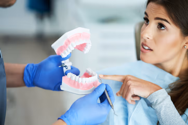 Dentist wearing blue gloves showing a dental model to a female patient who points at it during consultation.