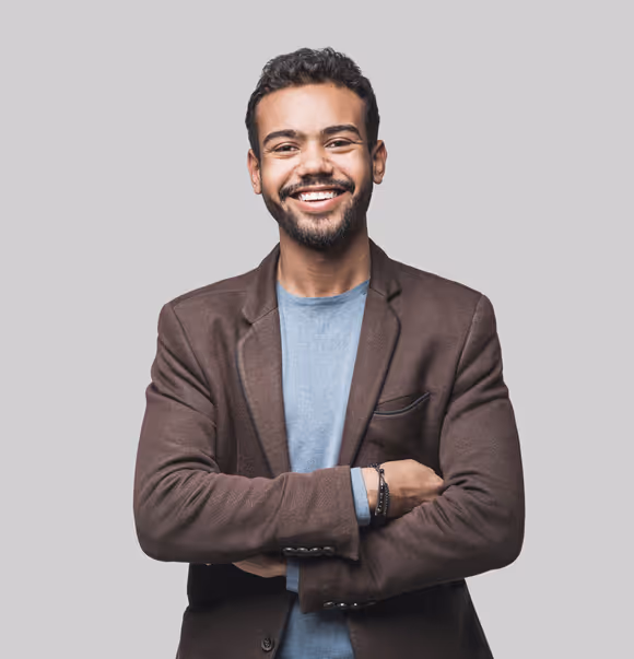 Smiling young man with curly hair and beard wearing a brown blazer and light blue shirt with arms crossed against a gray background.