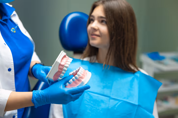 Dentist wearing blue gloves holding a dental model while a young patient with a blue bib looks on in a dental clinic.