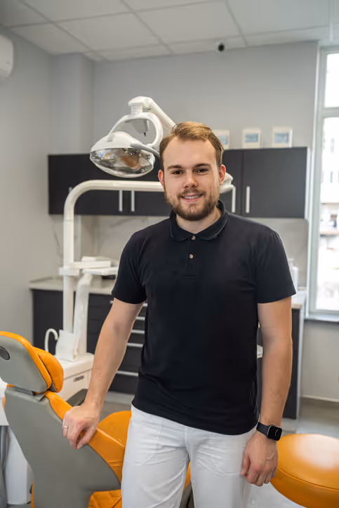 Young male dentist standing next to an orange dental chair in a modern dental clinic.