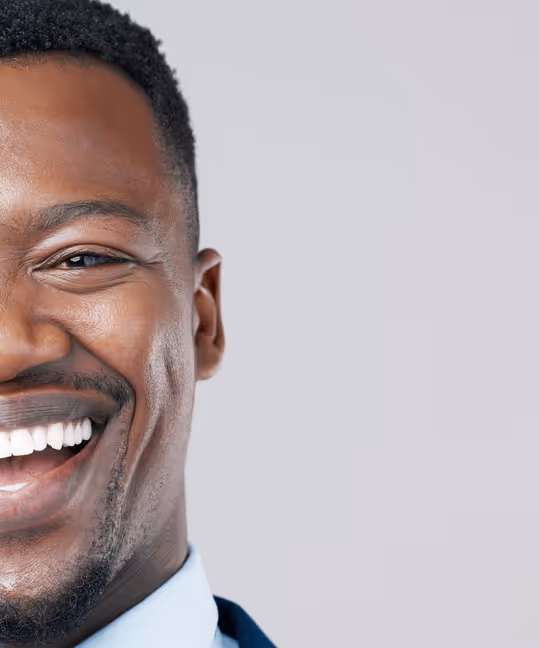 Close-up of a smiling man with dark skin, short curly hair, and a trimmed beard against a light gray background.