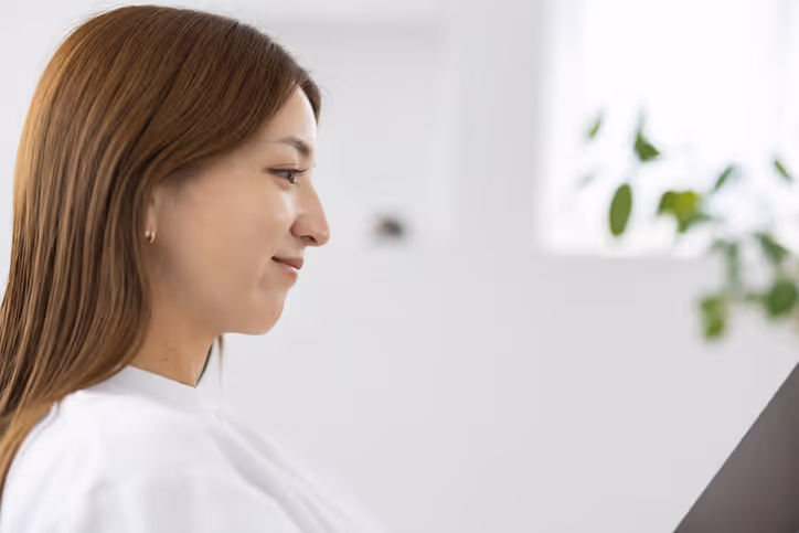 Smiling woman with long brown hair sitting in a white room looking at a black tablet or device.