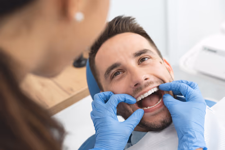 Dentist wearing blue gloves examining the open mouth and teeth of a smiling male patient reclined in a dental chair.
