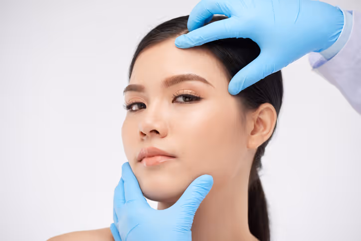 Close-up of a woman’s face being examined by hands wearing blue medical gloves against a white background.