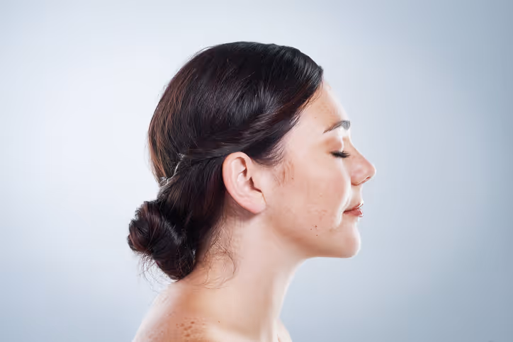 Profile view of a woman with dark hair in a low bun and eyes closed against a light gray background.
