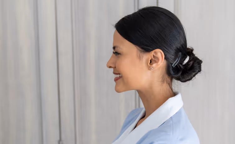Profile of a woman with dark hair tied in a bun, smiling and wearing a light blue blazer over a white shirt against a gray background.