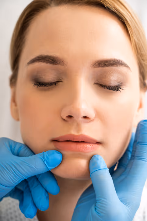 Close-up of a woman with closed eyes having her chin and jaw examined by hands wearing blue medical gloves.