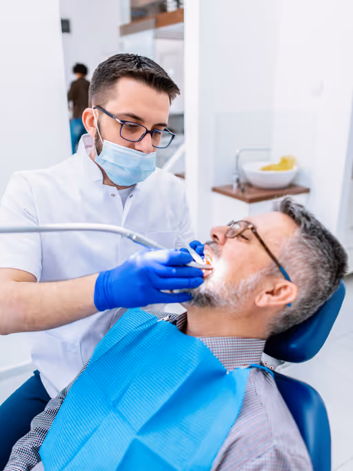 Dentist wearing a white coat, mask, and gloves examining a male patient with glasses reclining in a dental chair.
