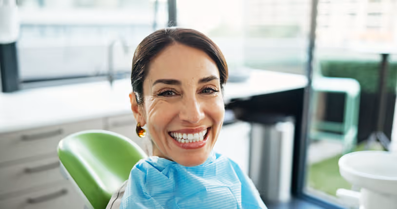 Smiling woman sitting in a dental chair wearing a blue dental bib in a bright dental office.