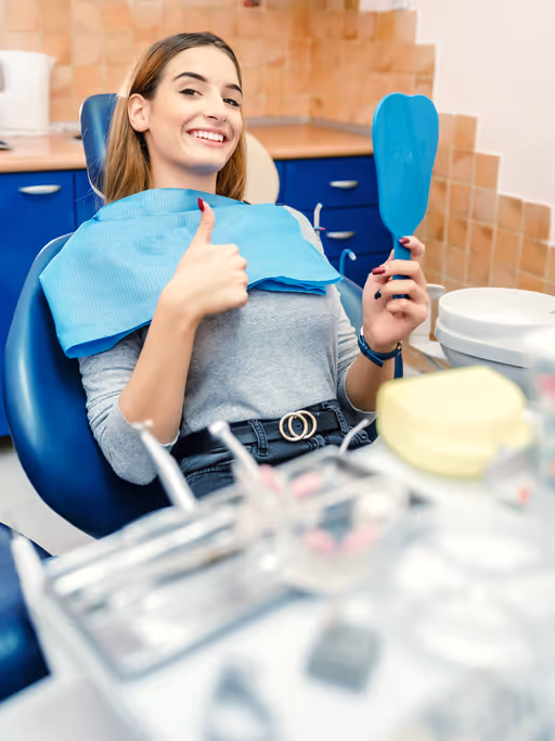 Smiling woman in dental chair giving thumbs up and holding a mirror in a dental office.