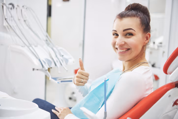 Smiling woman in a dental clinic giving a thumbs-up while sitting in a dental chair with a bib on.