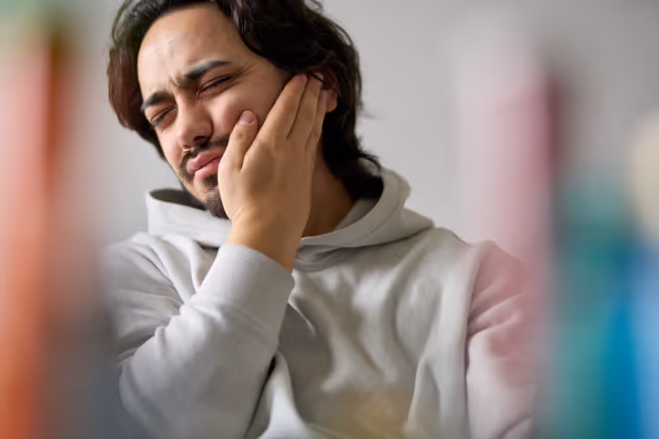 Young man in a gray hoodie holding his cheek in pain, indicating a toothache or jaw discomfort.