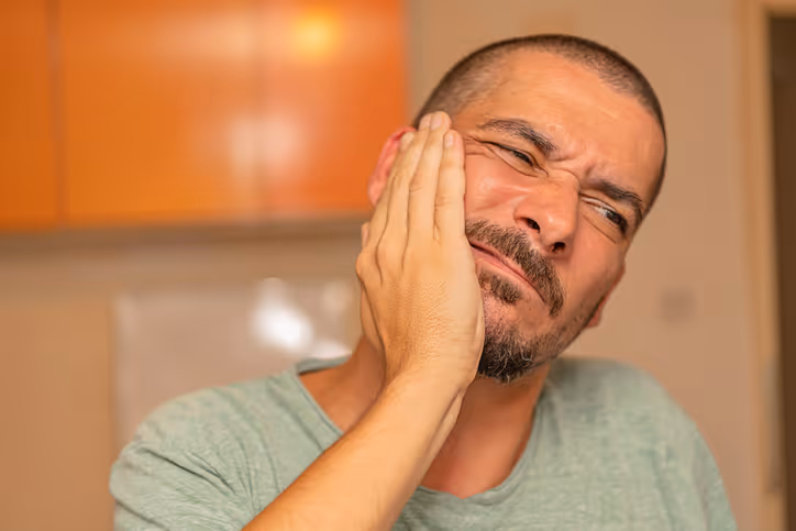Man with a beard and short hair wearing a green shirt holding his cheek in pain, suggesting a toothache.
