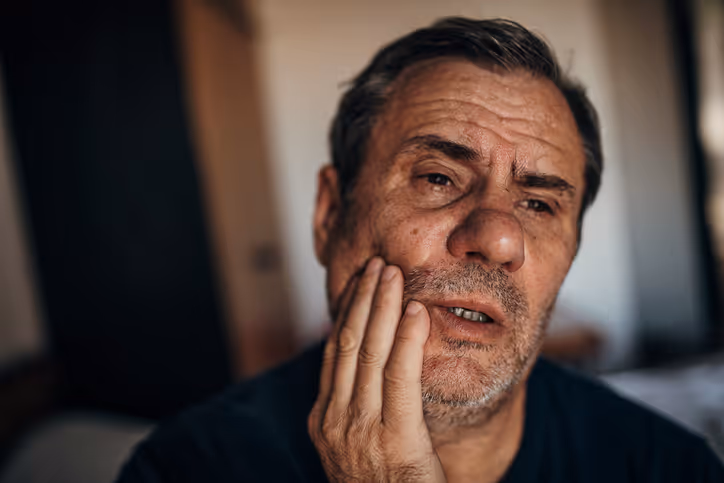 Middle-aged man with graying hair and beard touching his cheek, appearing to have tooth pain.