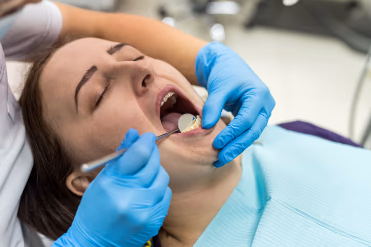 Dentist wearing blue gloves examining a young woman's open mouth with dental instruments during a checkup.