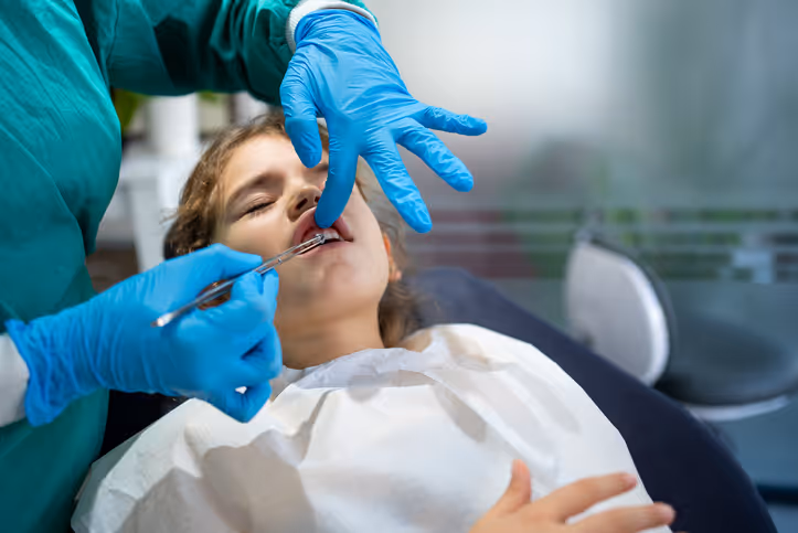 Dentist wearing blue gloves performing a dental procedure on a young girl lying back in a dental chair.