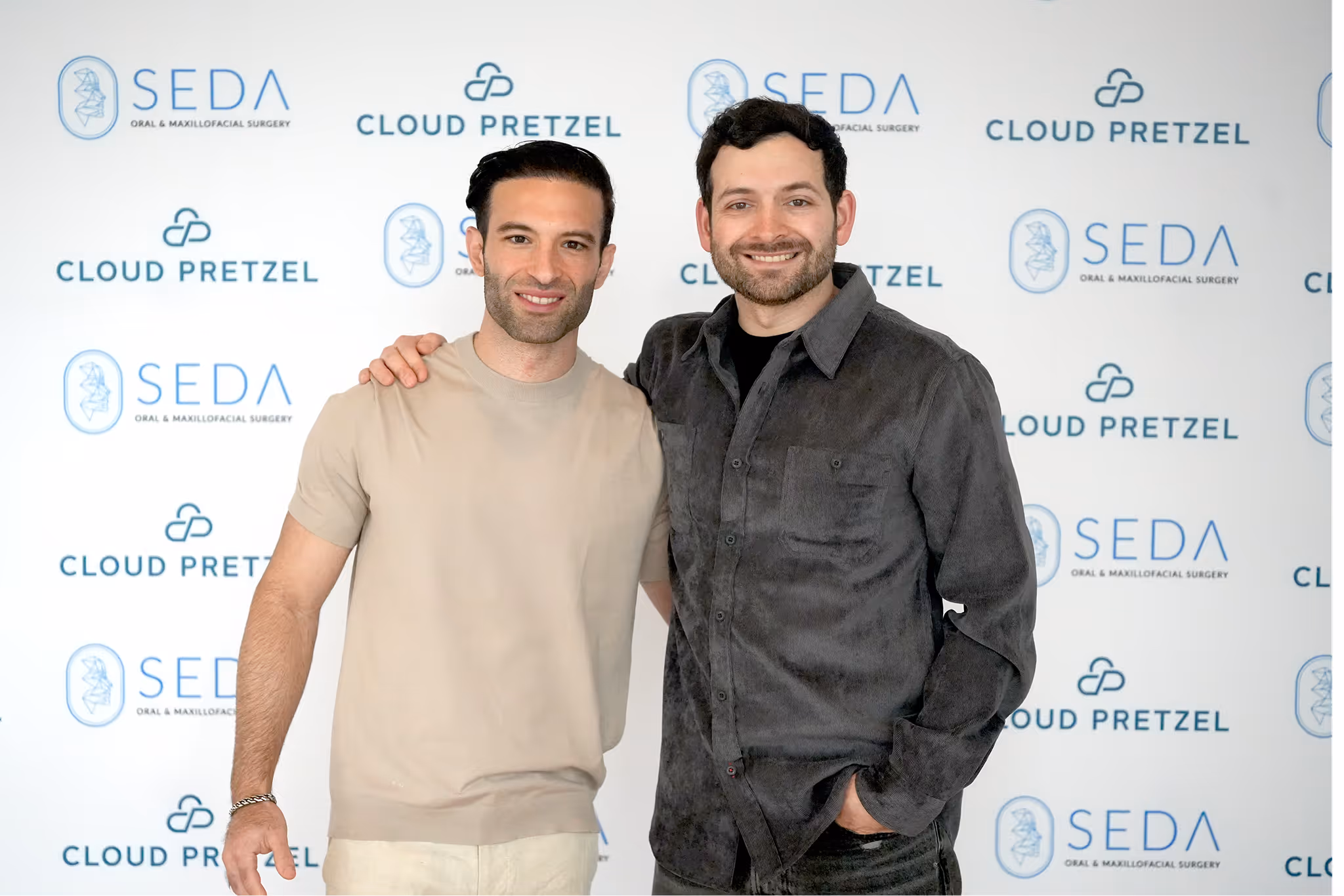 Two men standing close, smiling with one man's arm around the other's shoulder, against a backdrop featuring logos for SEDA Oral & Maxillofacial Surgery and Cloud Pretzel.