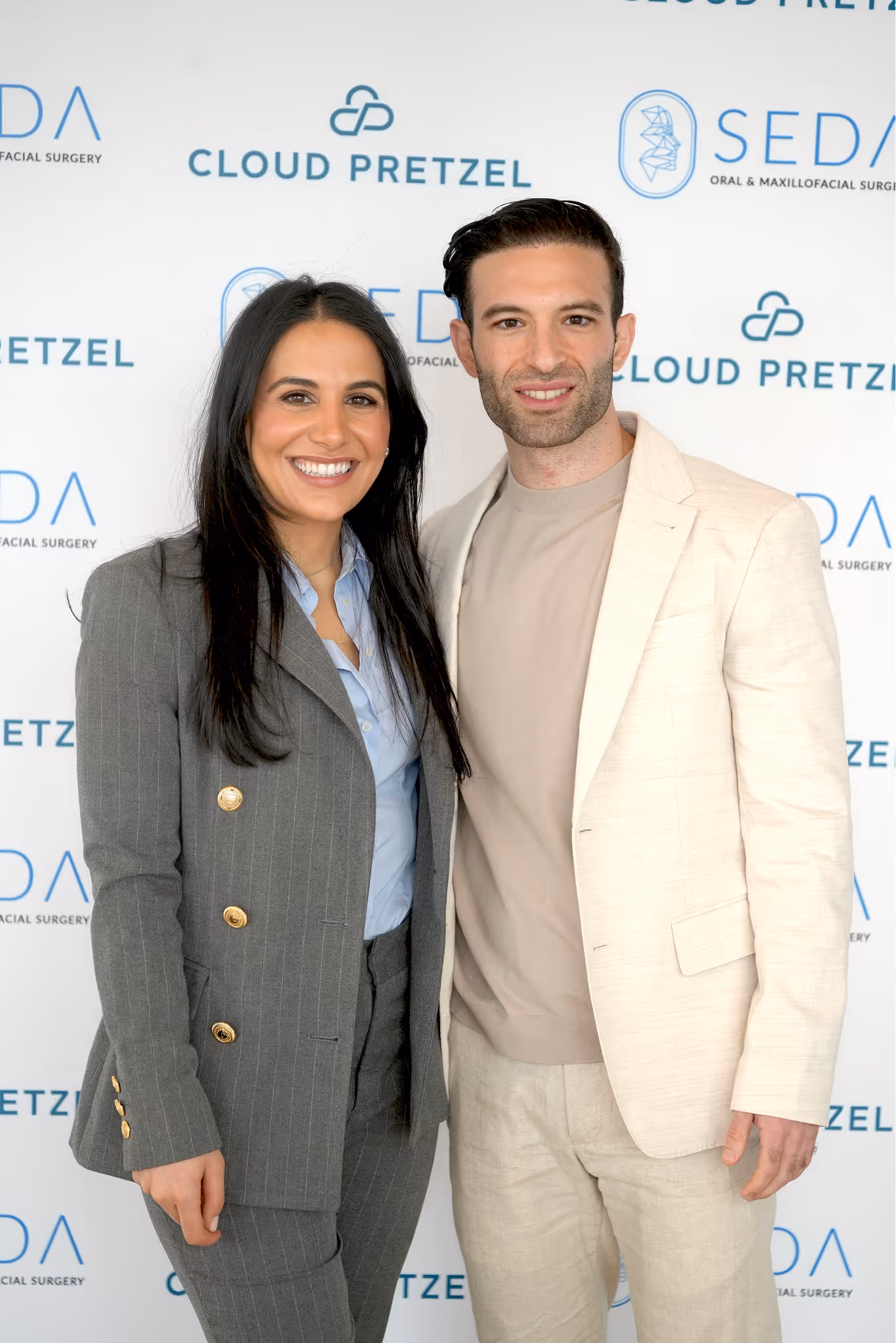 Smiling man and woman standing together in front of a white backdrop with Cloud Pretzel and Seda Oral & Maxillofacial Surgery logos.