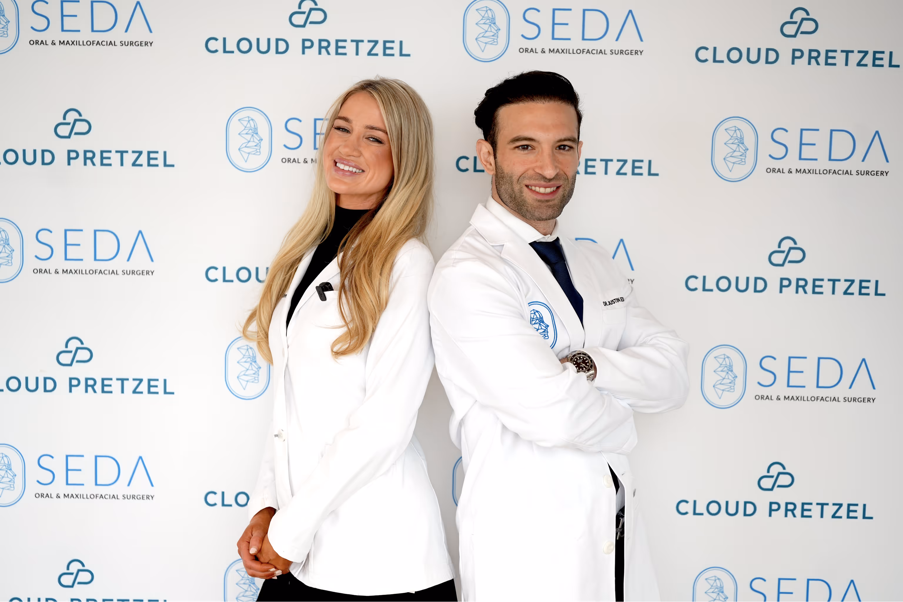 A smiling man and woman, both wearing white medical coats, standing back to back in front of a backdrop with SEDA Oral & Maxillofacial Surgery and Cloud Pretzel logos.