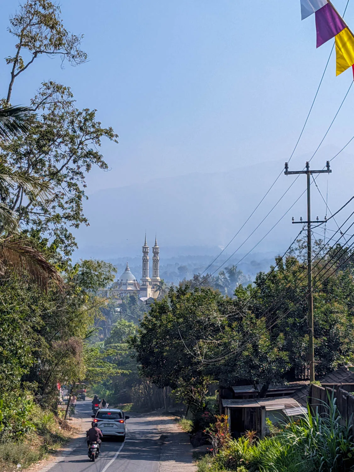 "Rural road in Central Lombok winding downhill towards a domed mosque with twin minarets, set against hazy hills