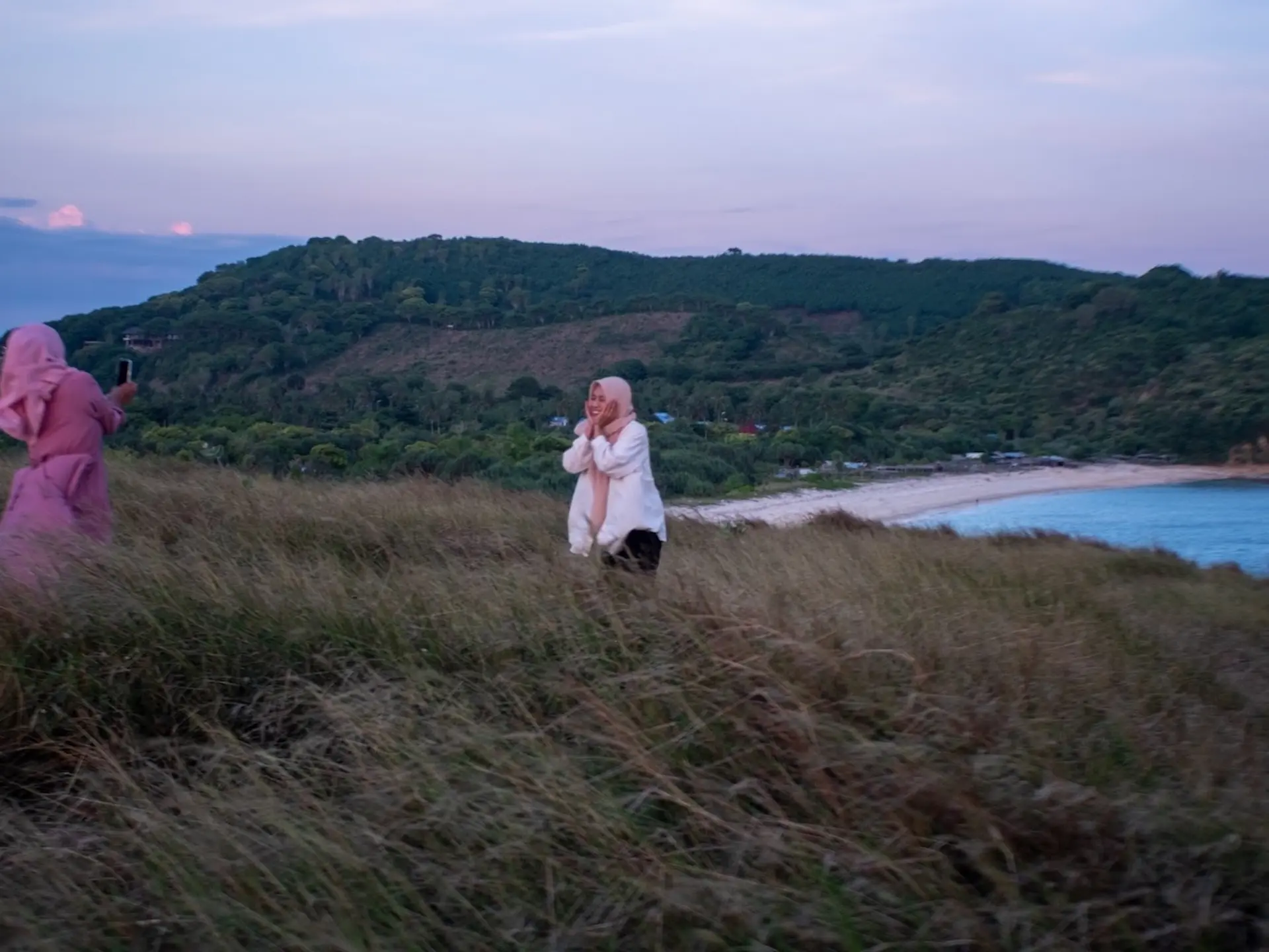 Woman posing on a grassy hill at Bukit Pedau , Lombok overlooking the sea at sunset with another women taking a picture