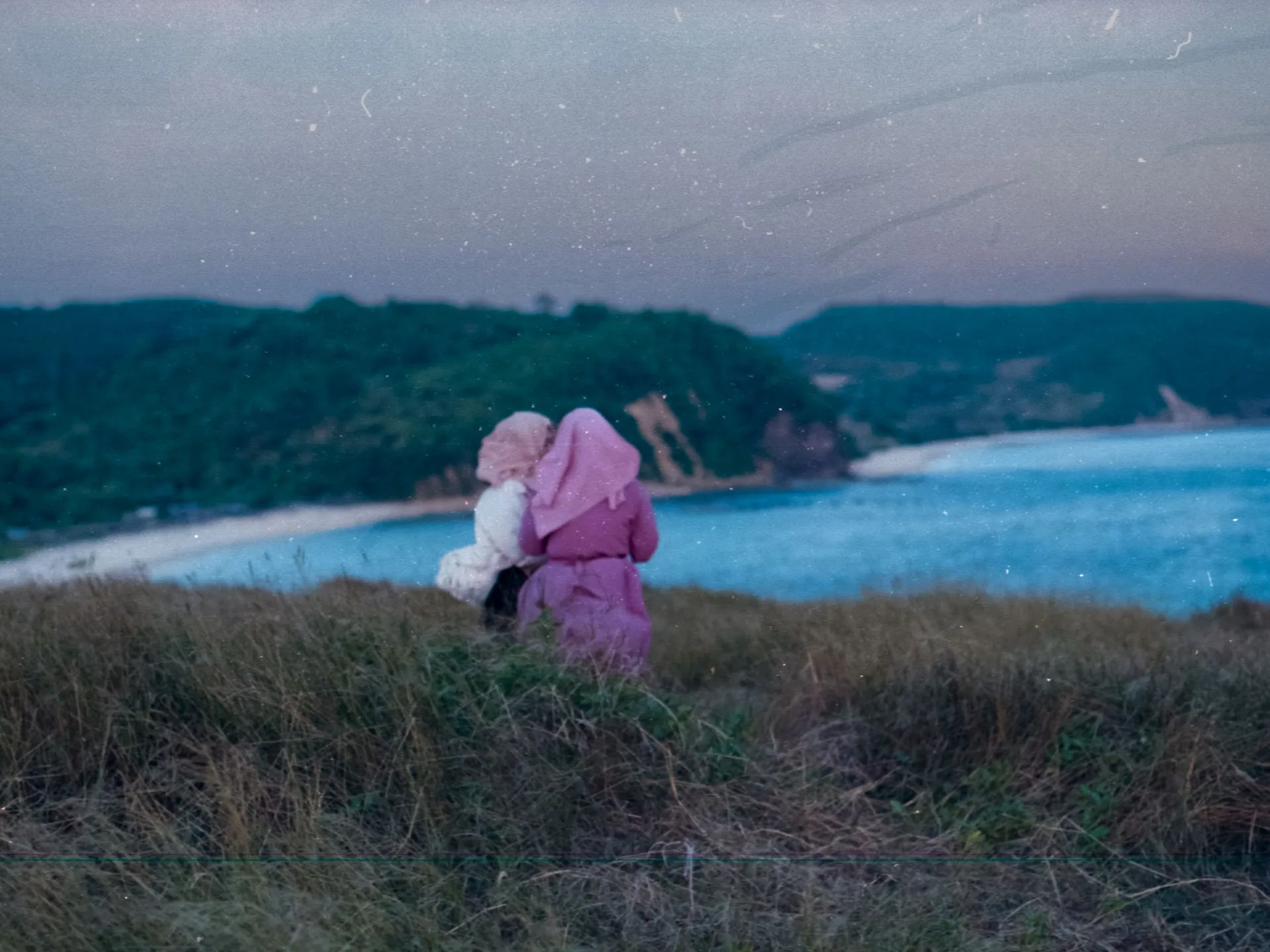 Woman posing on a grassy hill at Bukit Pedau, Lombok overlooking the sea at sunset with another woman taking a picture