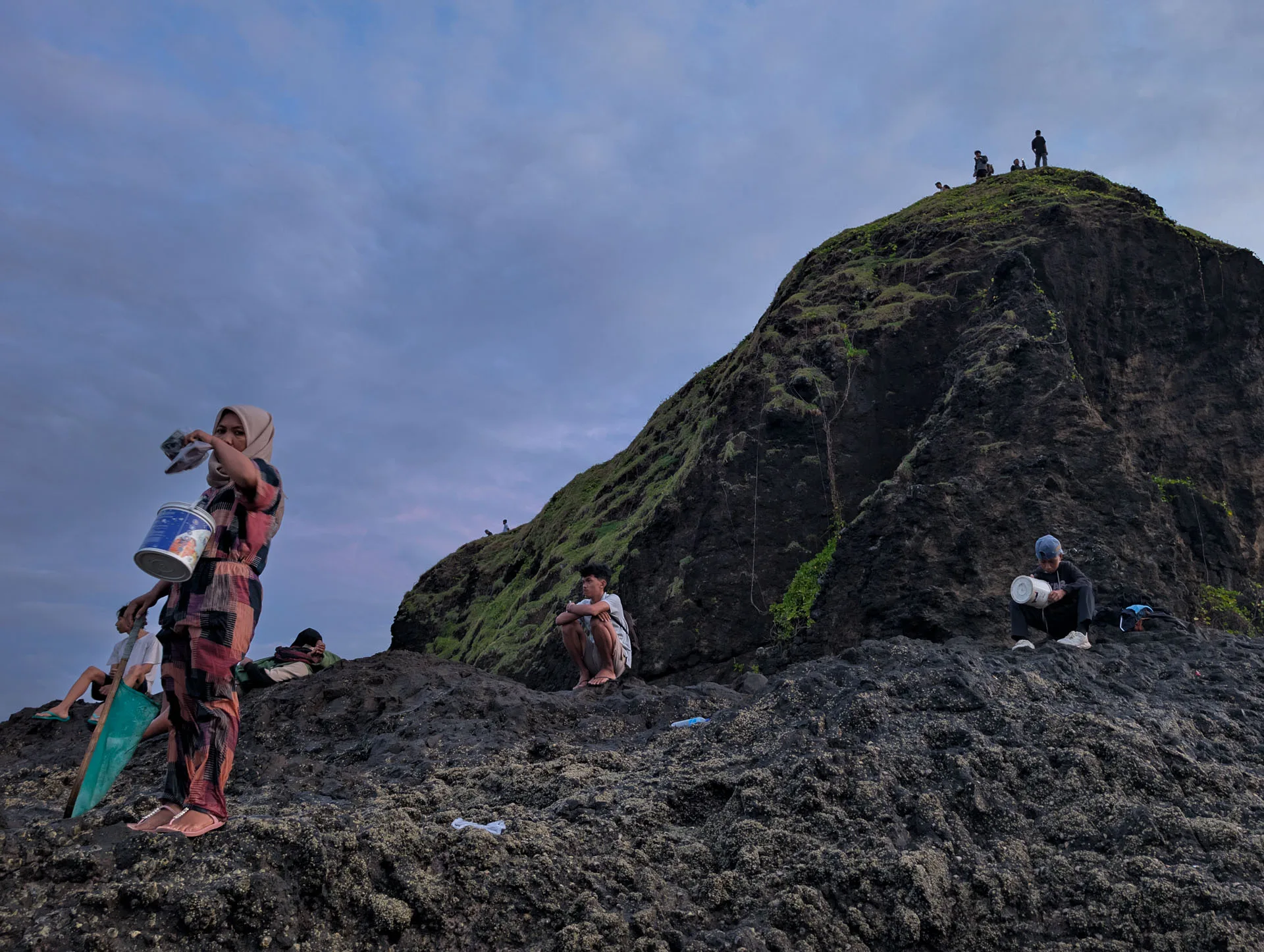 A woman holding a bucket and net stands on the rocks below a grass-topped headland at Seger beach, with people on its summit
