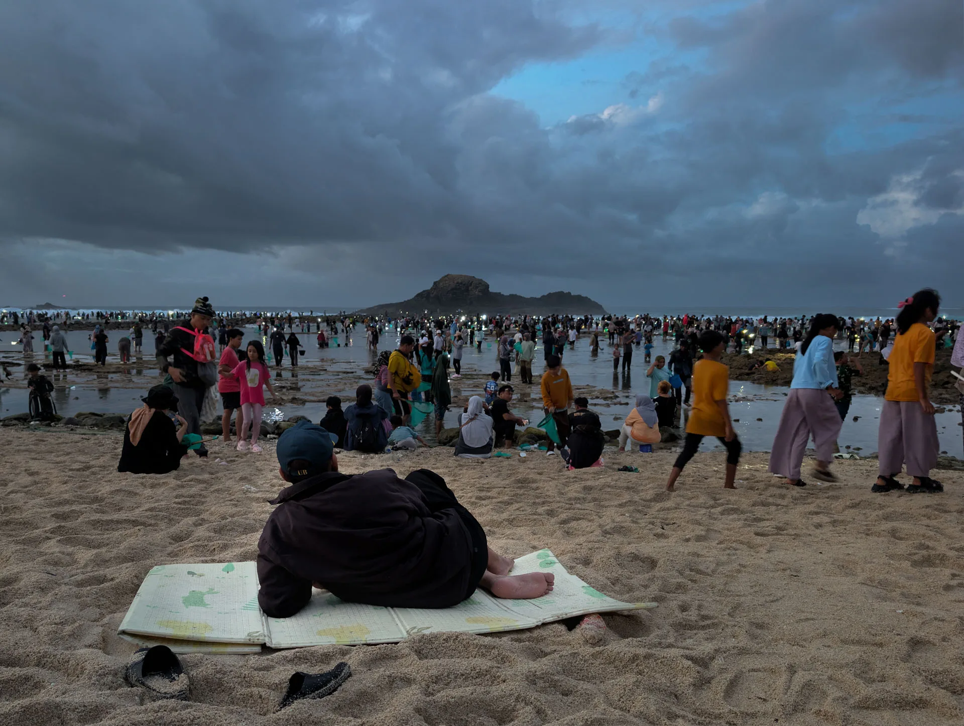 A man lies on a mat on the sand, watching the crowd in the tide pools at Seger beach after dawn