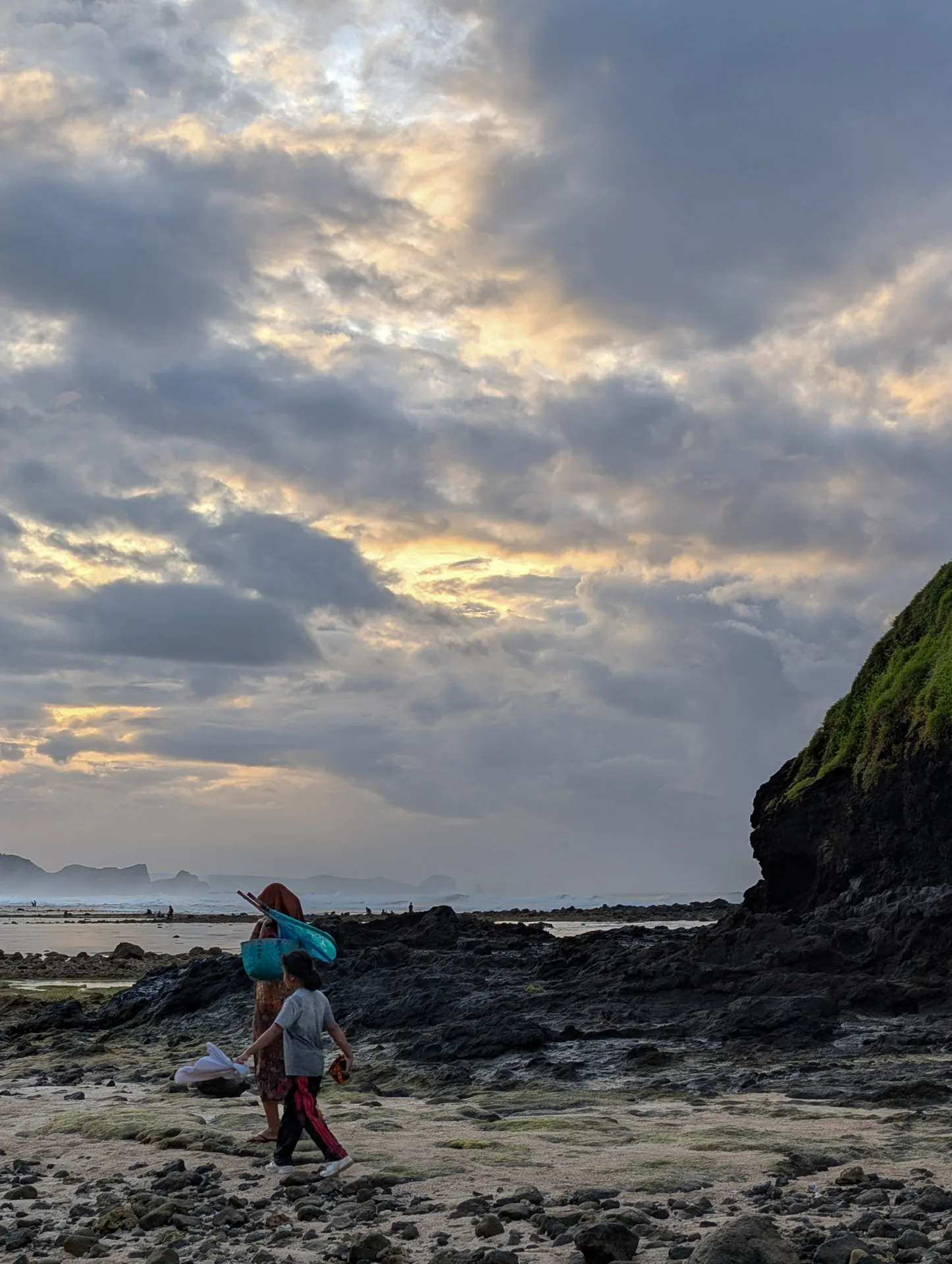 A woman and a girl carry a net and basket across the rocks at Seger beach, walking away from the sea