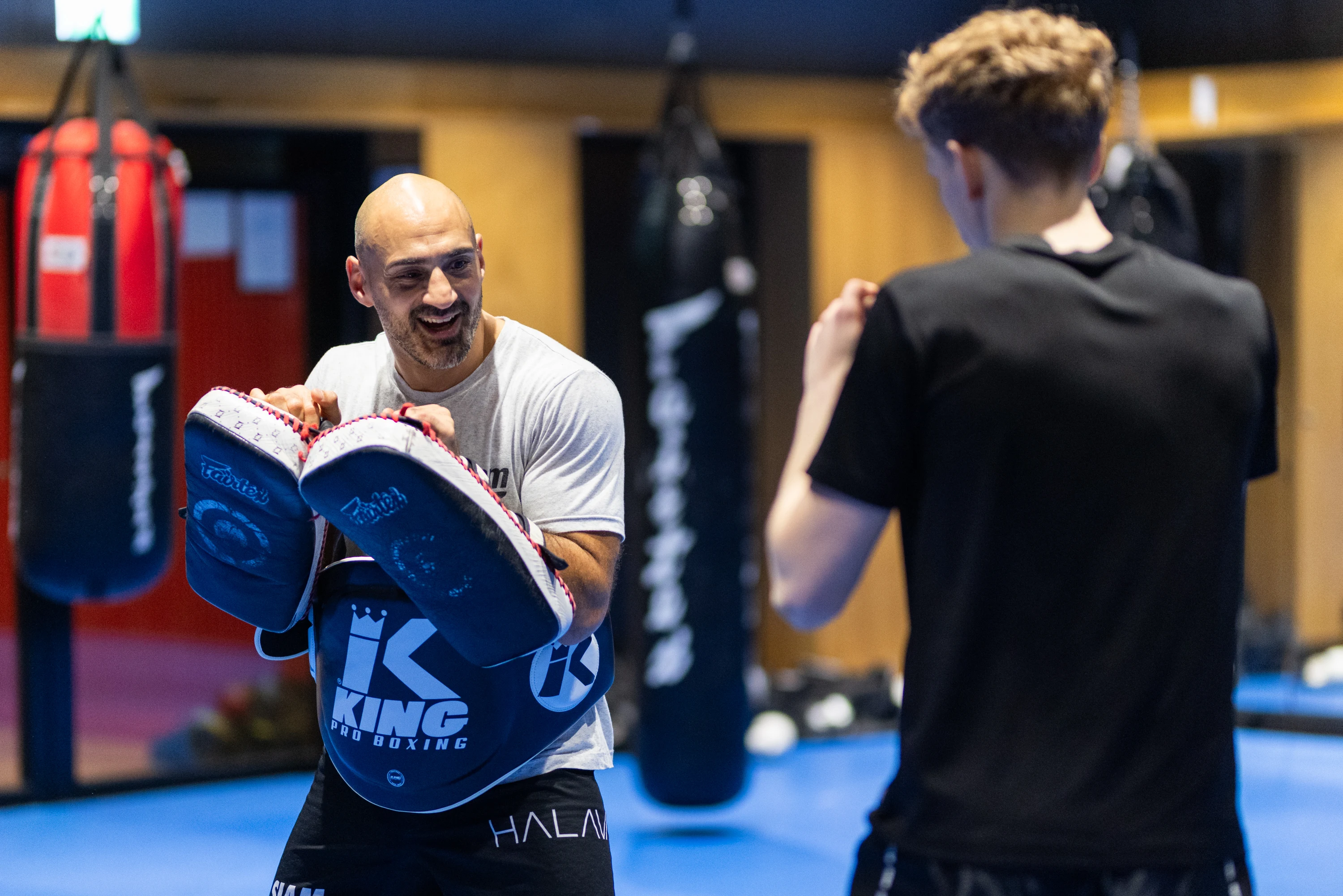 Boxing coach holding focus mitts smiling at a young boxer in a gym with punching bags in the background.