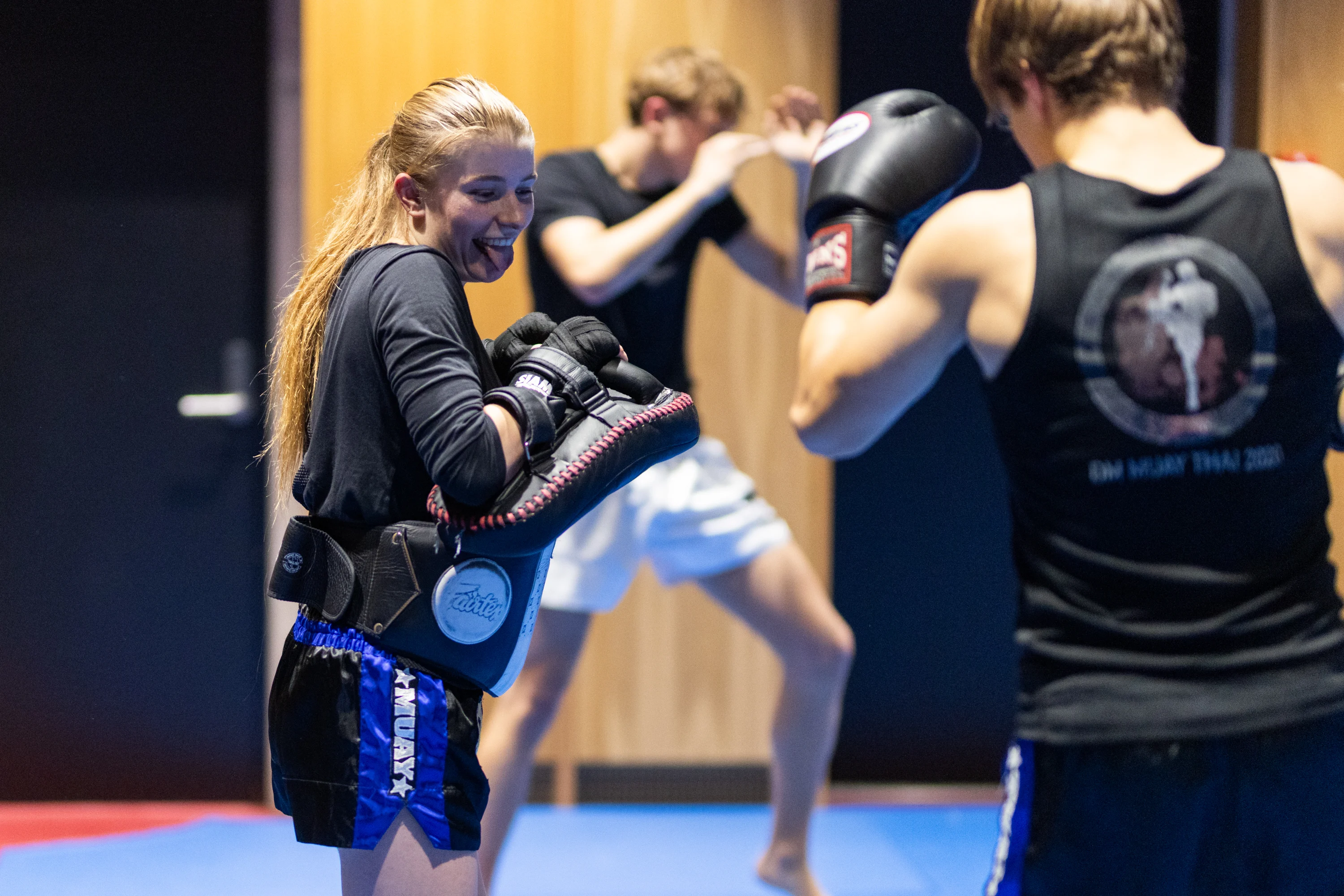 Two people sparring in a Muay Thai gym, with a female trainer holding pads and smiling while a male boxer prepares to punch.