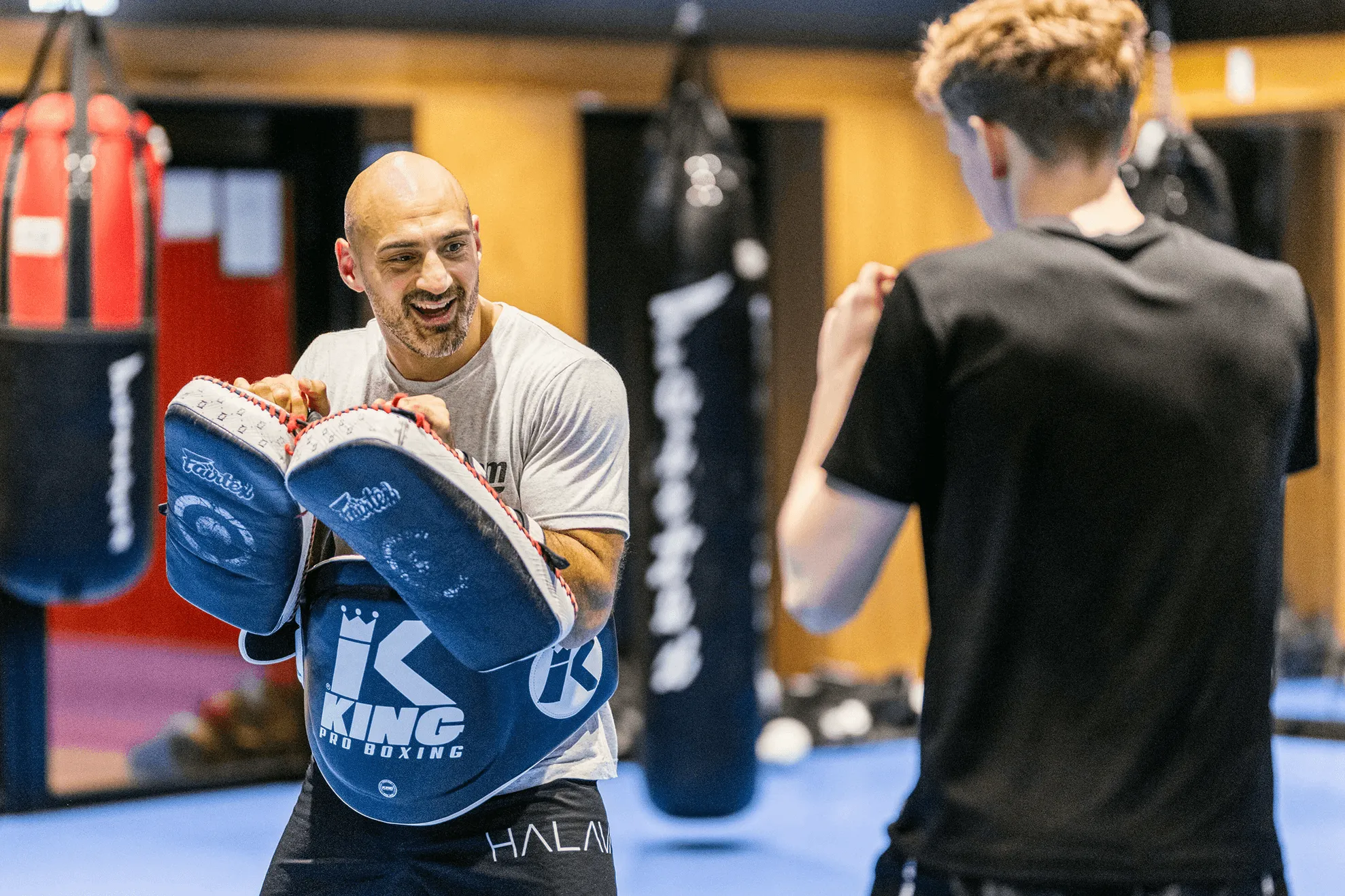 Boxing coach holding focus mitts for a young male boxer during training in a gym with punching bags.