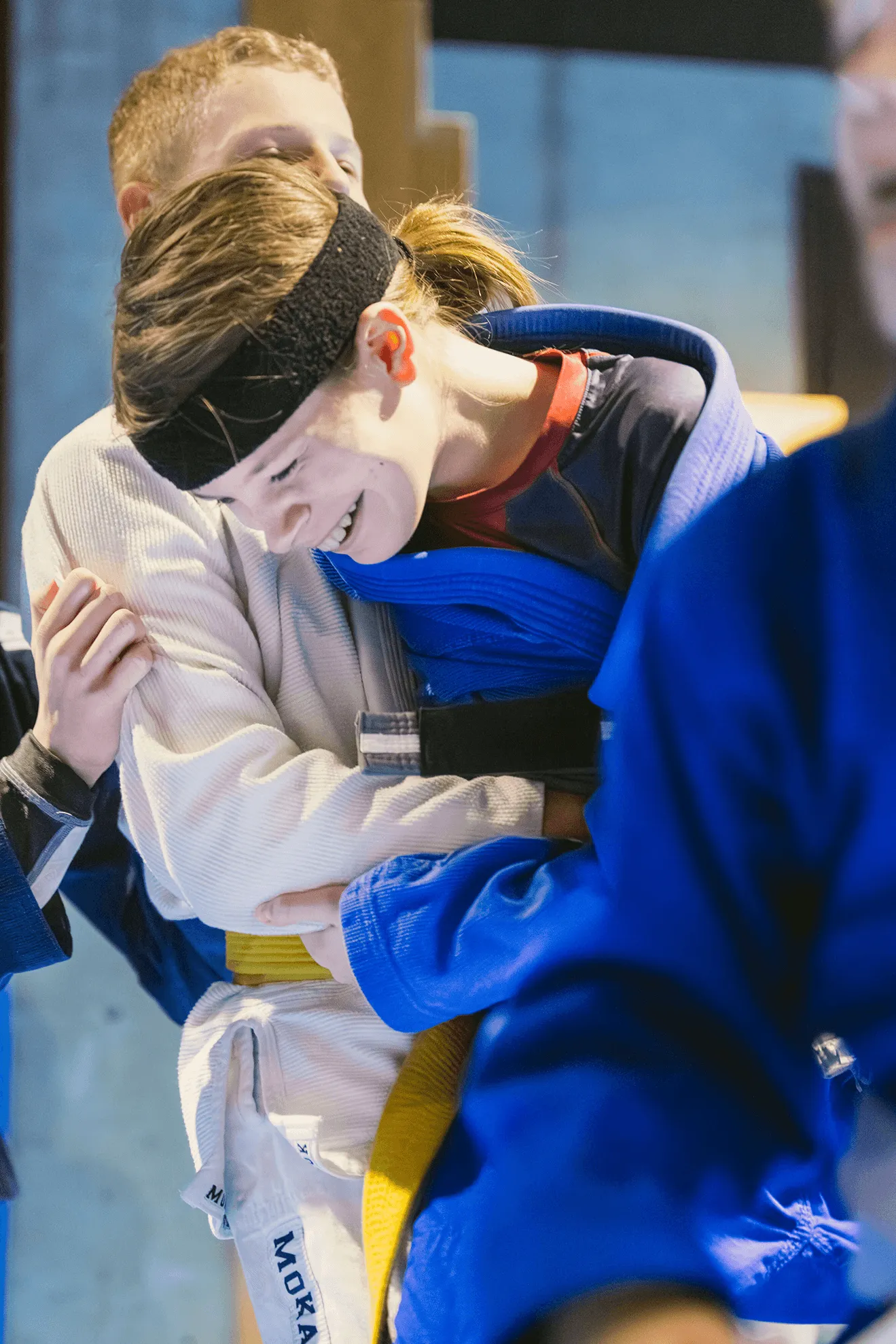 Two men engaged in grappling on a blue training mat inside a gym with large windows.