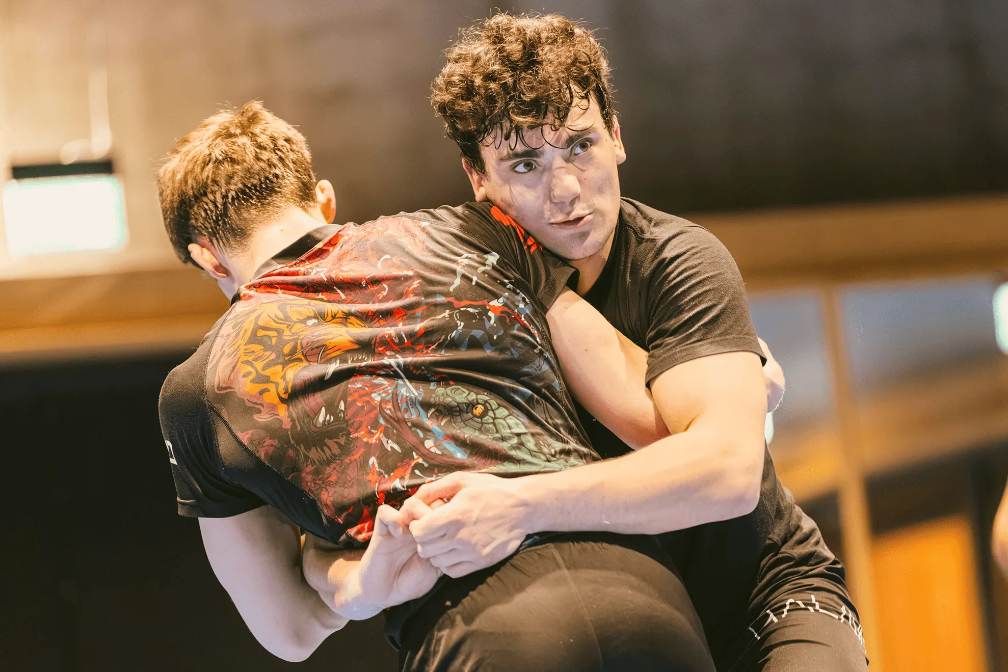 Two men practicing grappling on a blue mat in an indoor training gym.