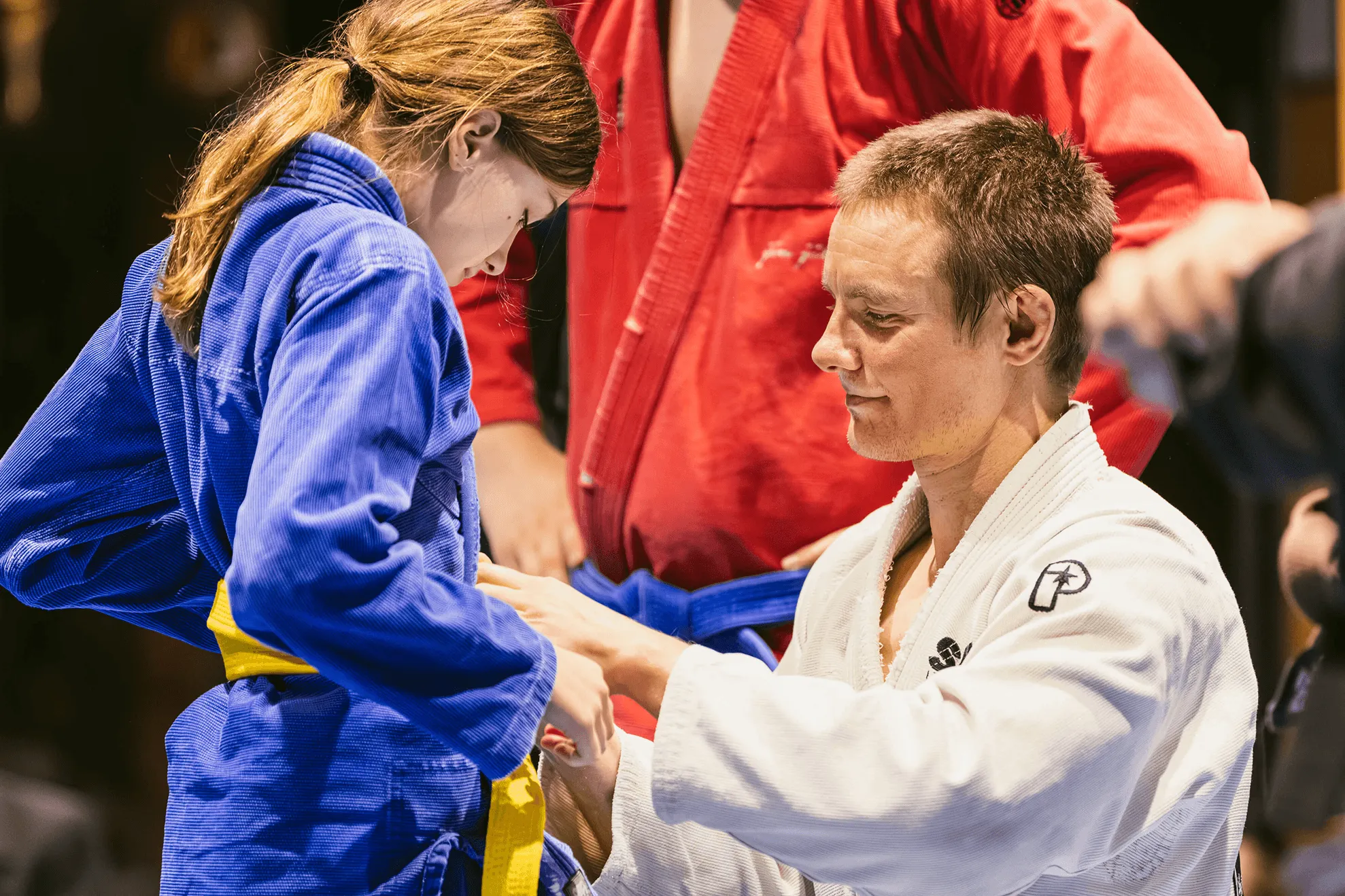 Man in white martial arts uniform tying yellow belt on young girl wearing blue martial arts uniform.