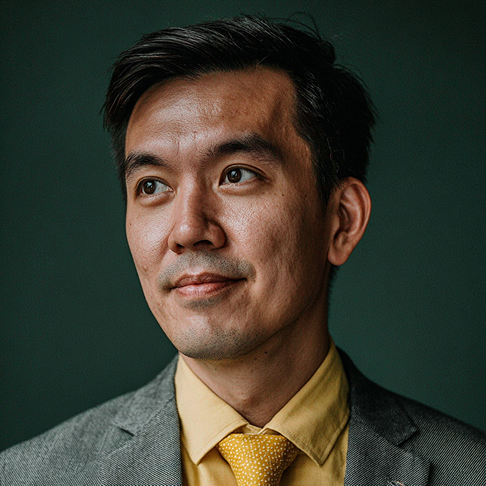 Portrait of a man wearing a gray suit, yellow shirt, and patterned yellow tie against a dark background.