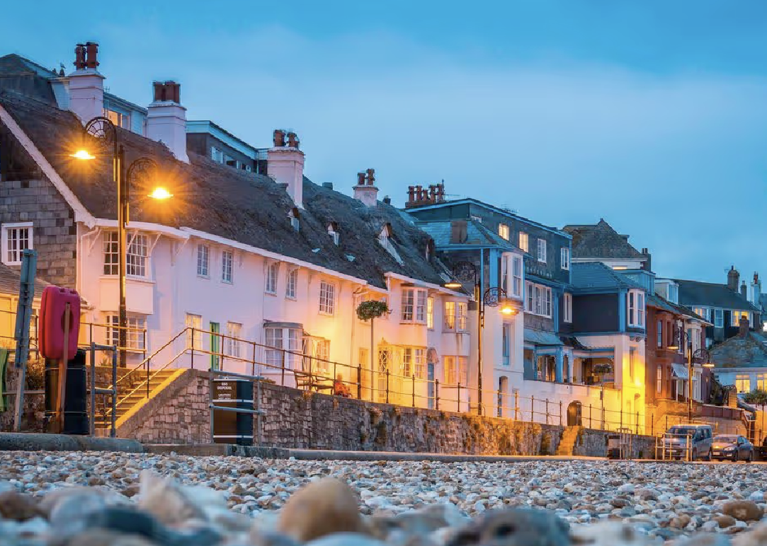 Pebble beach with lit street lamps and white cottages along a stone wall at dusk.