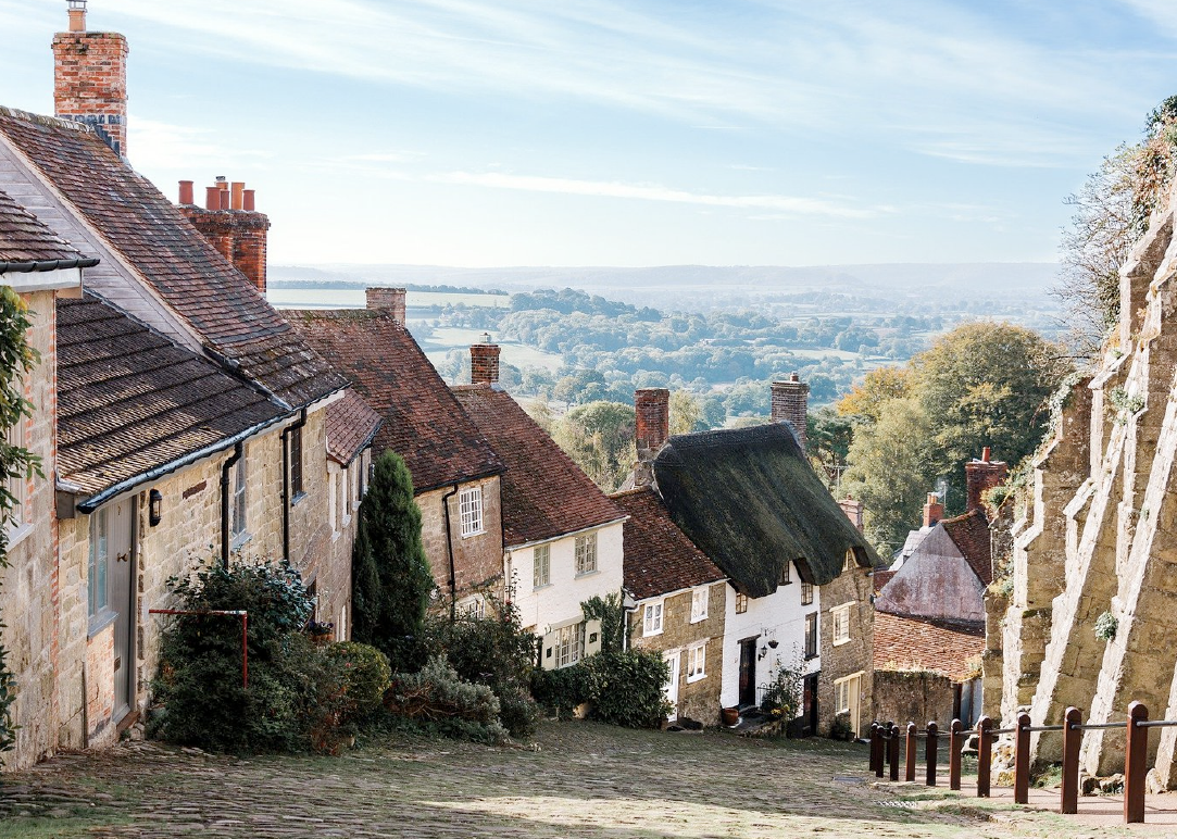 Historic stone doreset cottages with thatched and tiled roofs on a steep cobblestone street overlooking countryside.