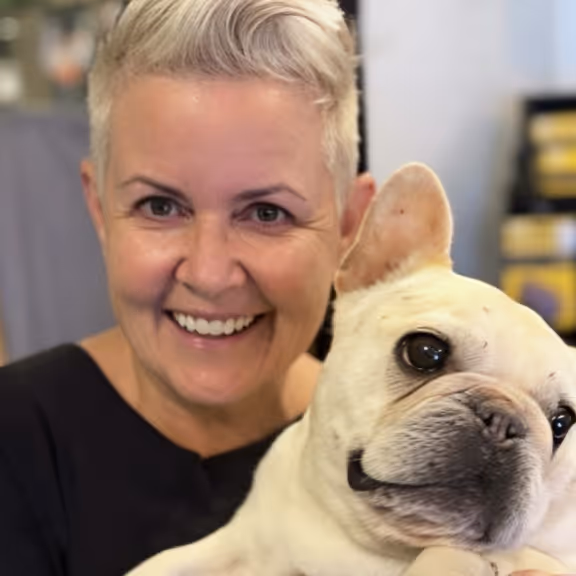 Smiling woman with short gray hair holding a white French Bulldog close to her face.