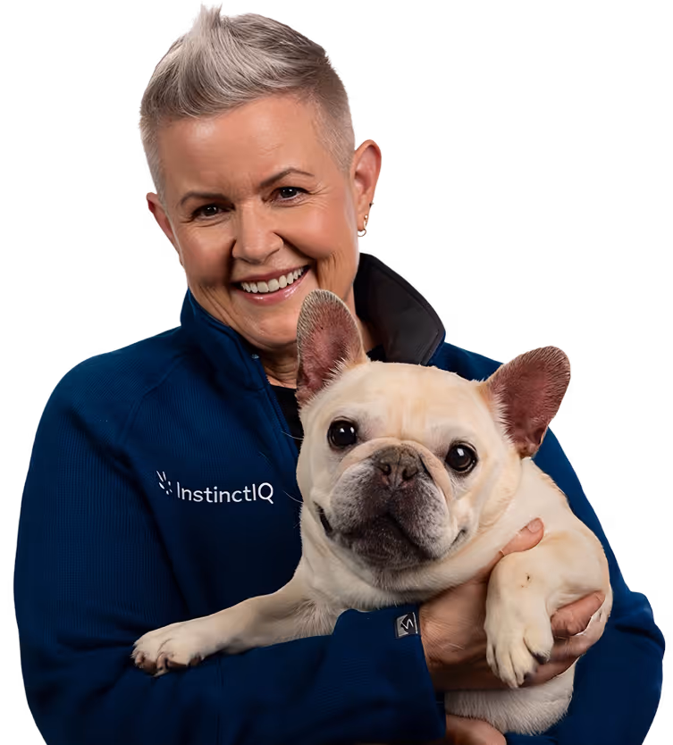Smiling woman in navy InstinctIQ jacket holding a cream-colored French bulldog against black background.