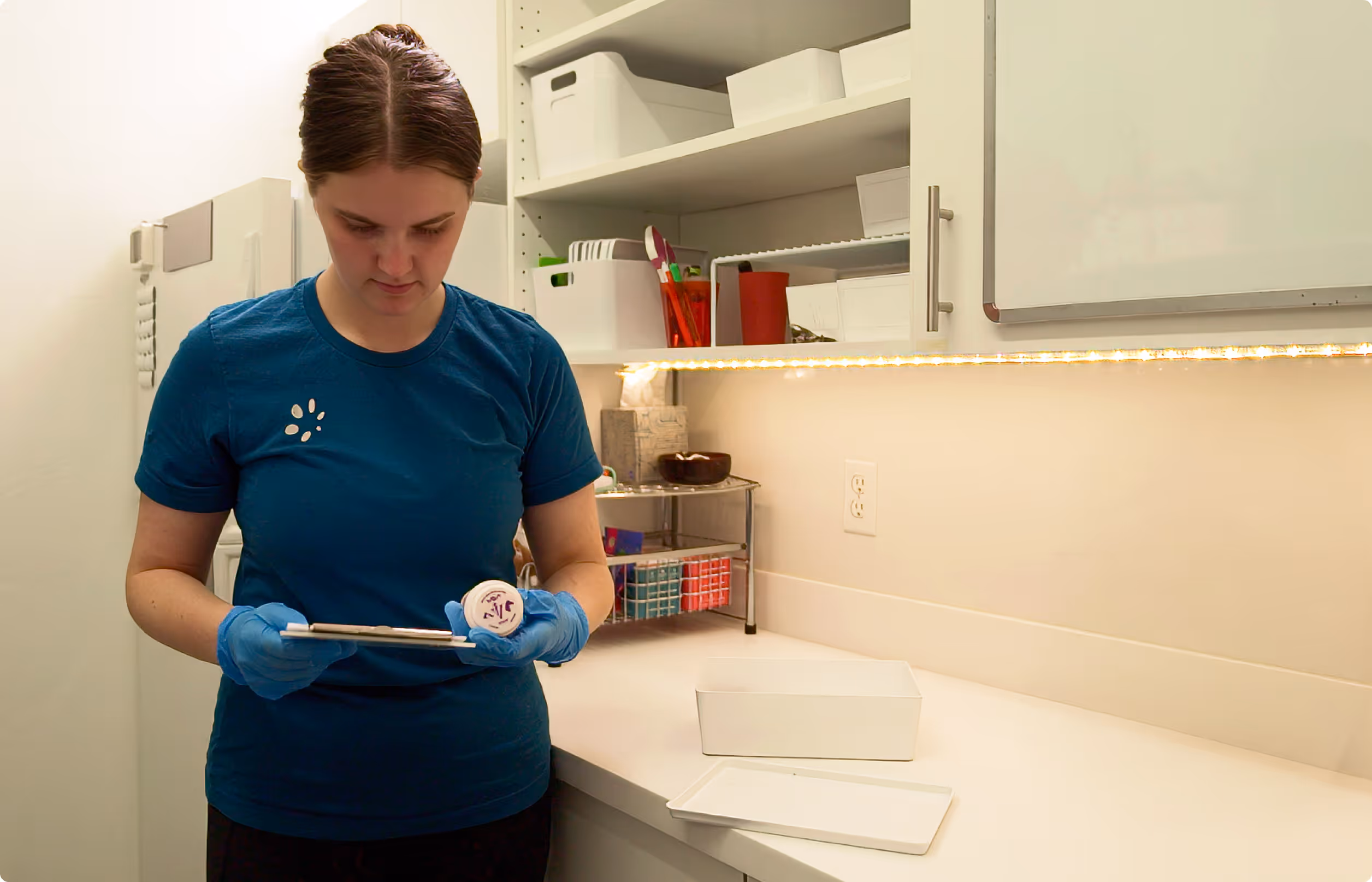 Person wearing blue gloves holds a prescription bottle and tablet in a clean, organized pharmacy room.