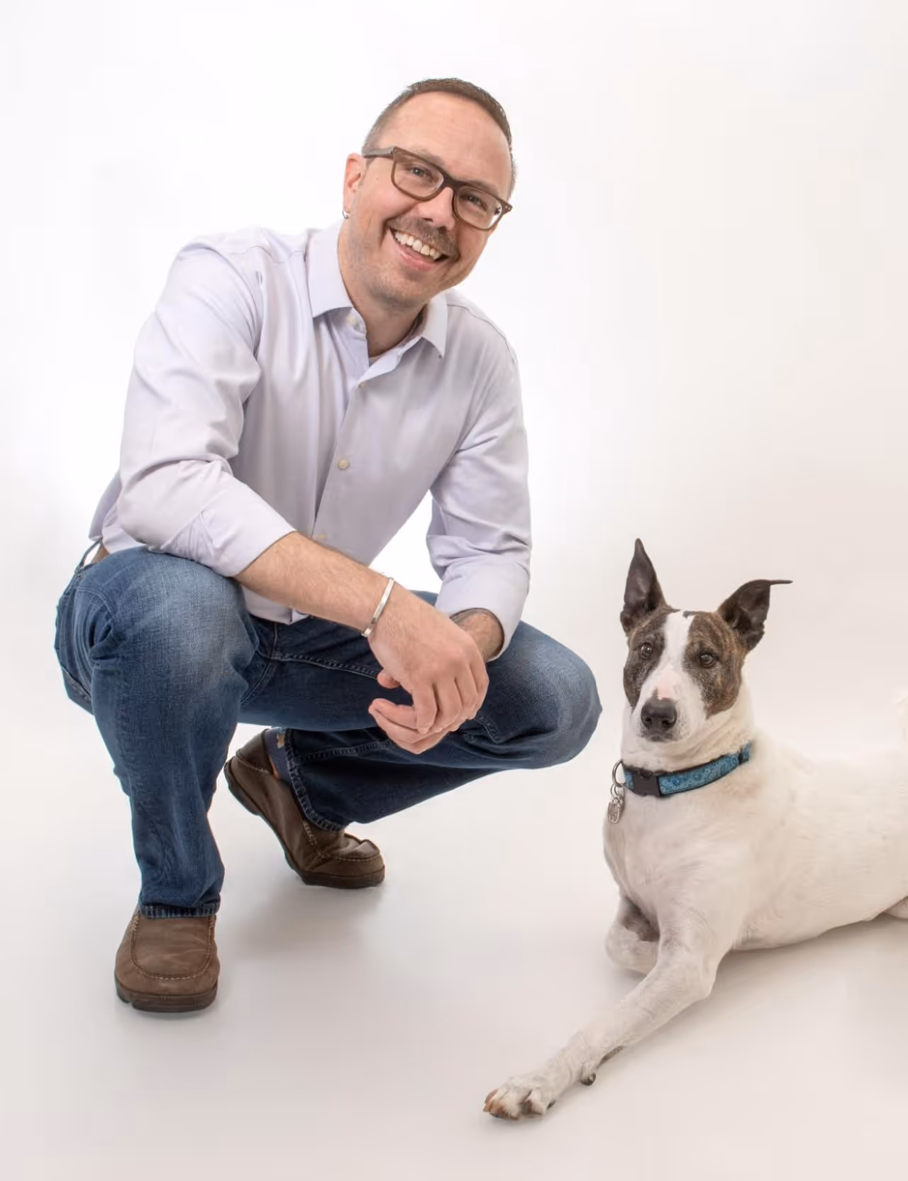 Smiling man in glasses squats next to a white dog with a blue collar on a white background.