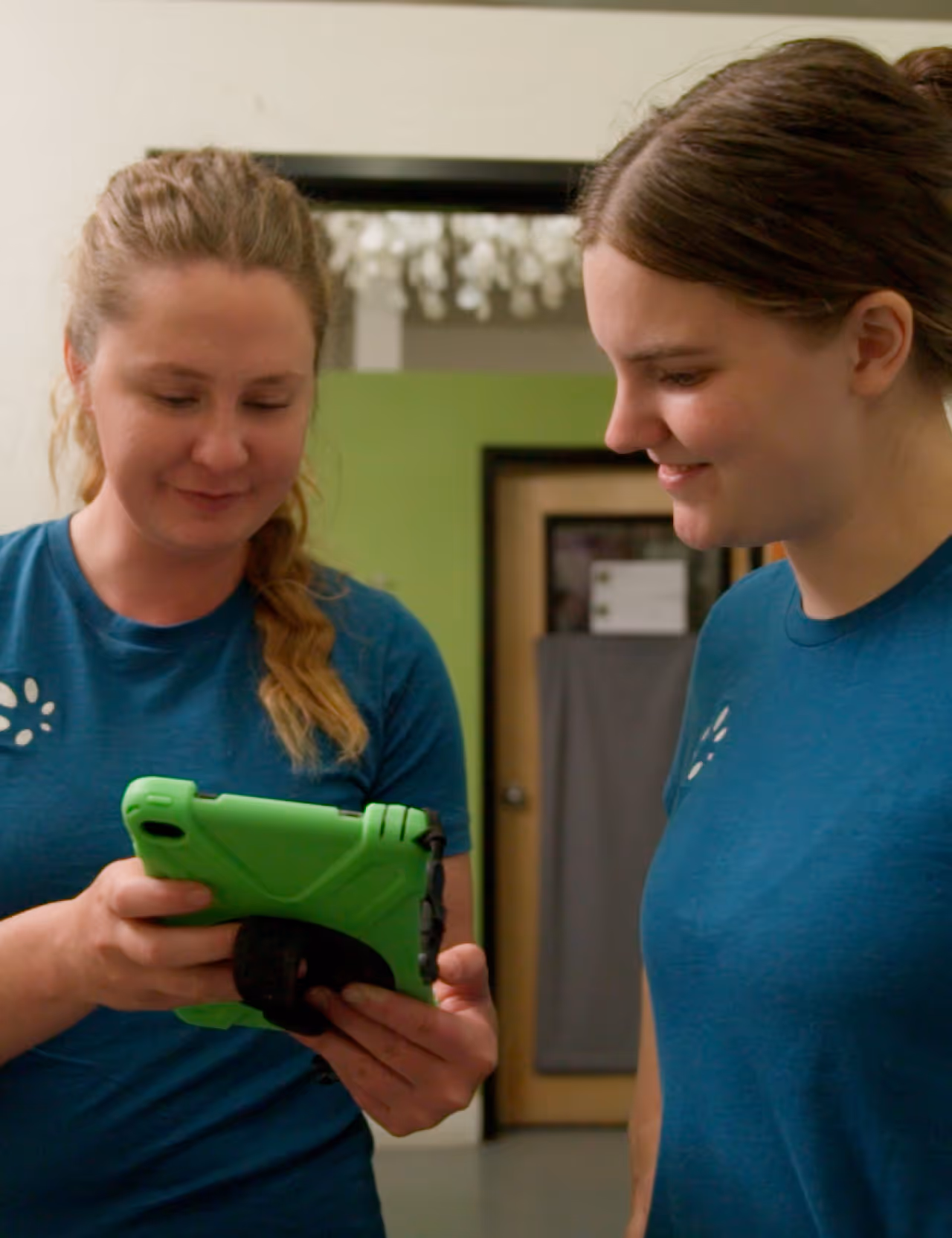 Two women in blue shirts looking at a green tablet in an indoor setting.