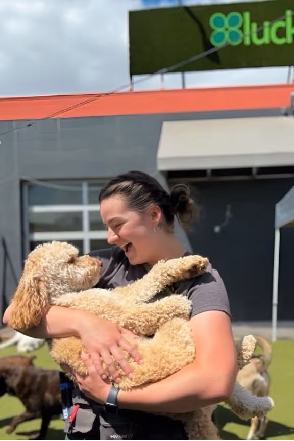 Woman smiling happily while holding a large curly-haired dog outdoors at a pet care facility.