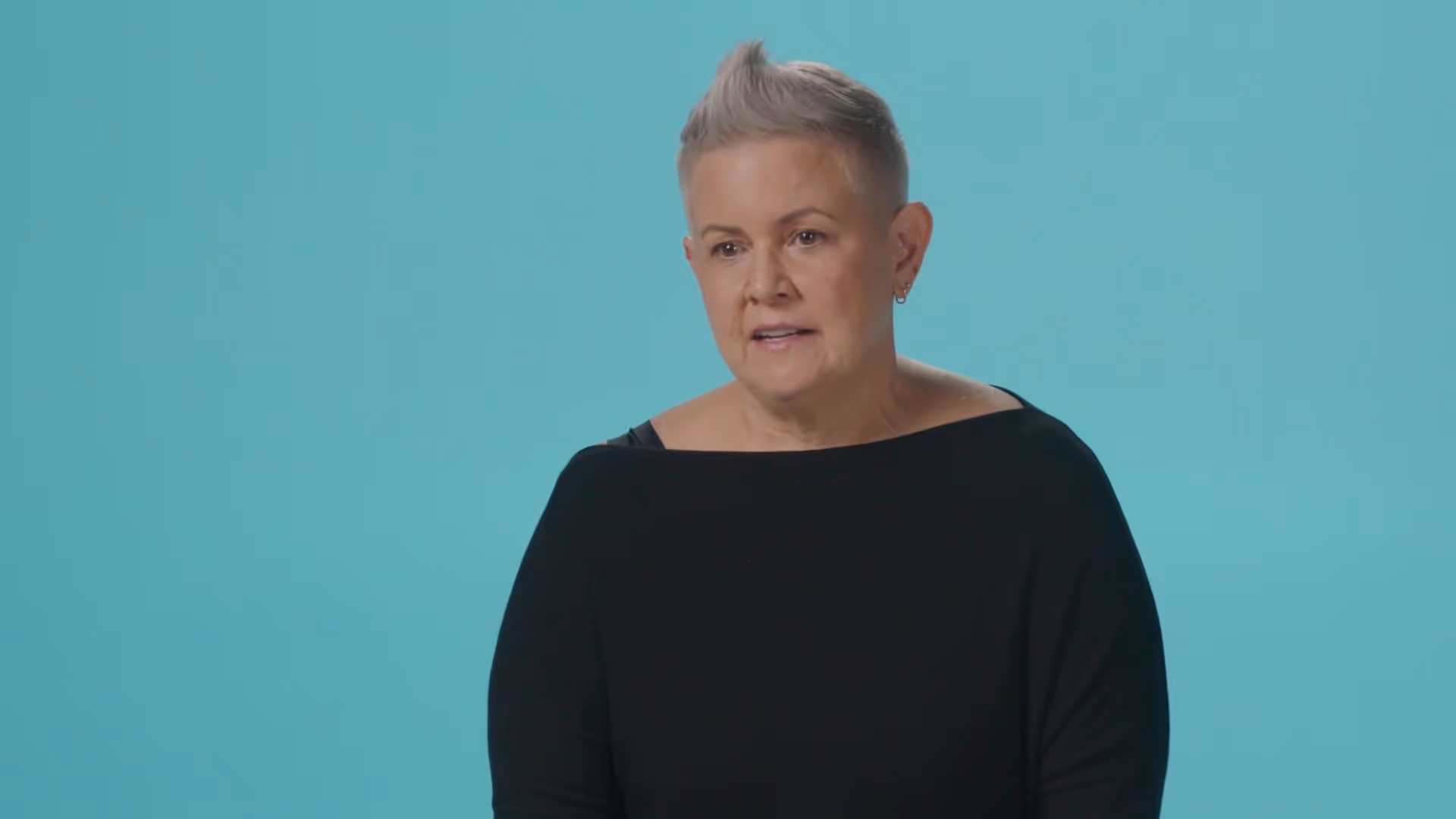 Middle-aged woman with short gray hair in a black top speaking against a teal background.