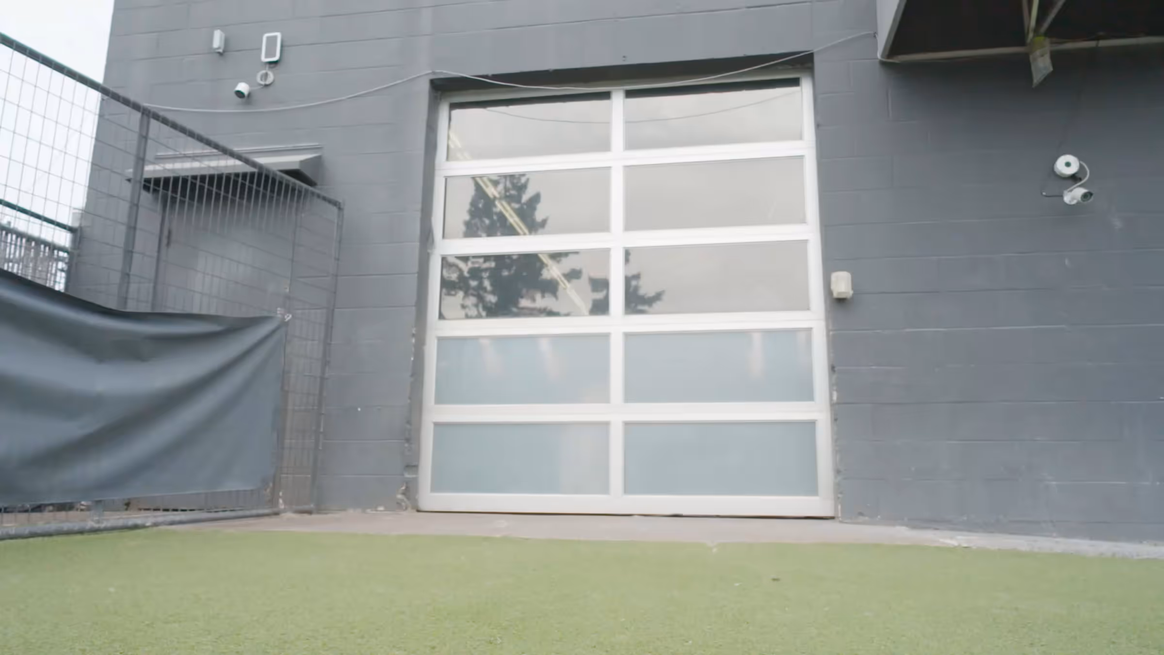 White paneled garage door on gray building wall with security cameras and green artificial turf in front.