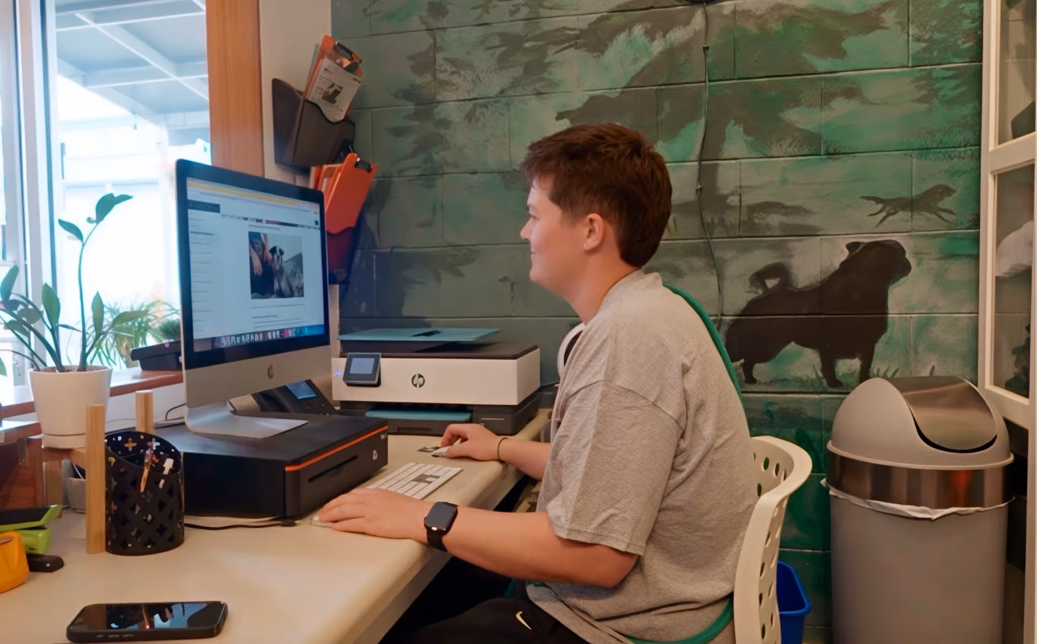 Person sitting at desk using desktop computer with dog mural on wall and office supplies nearby.