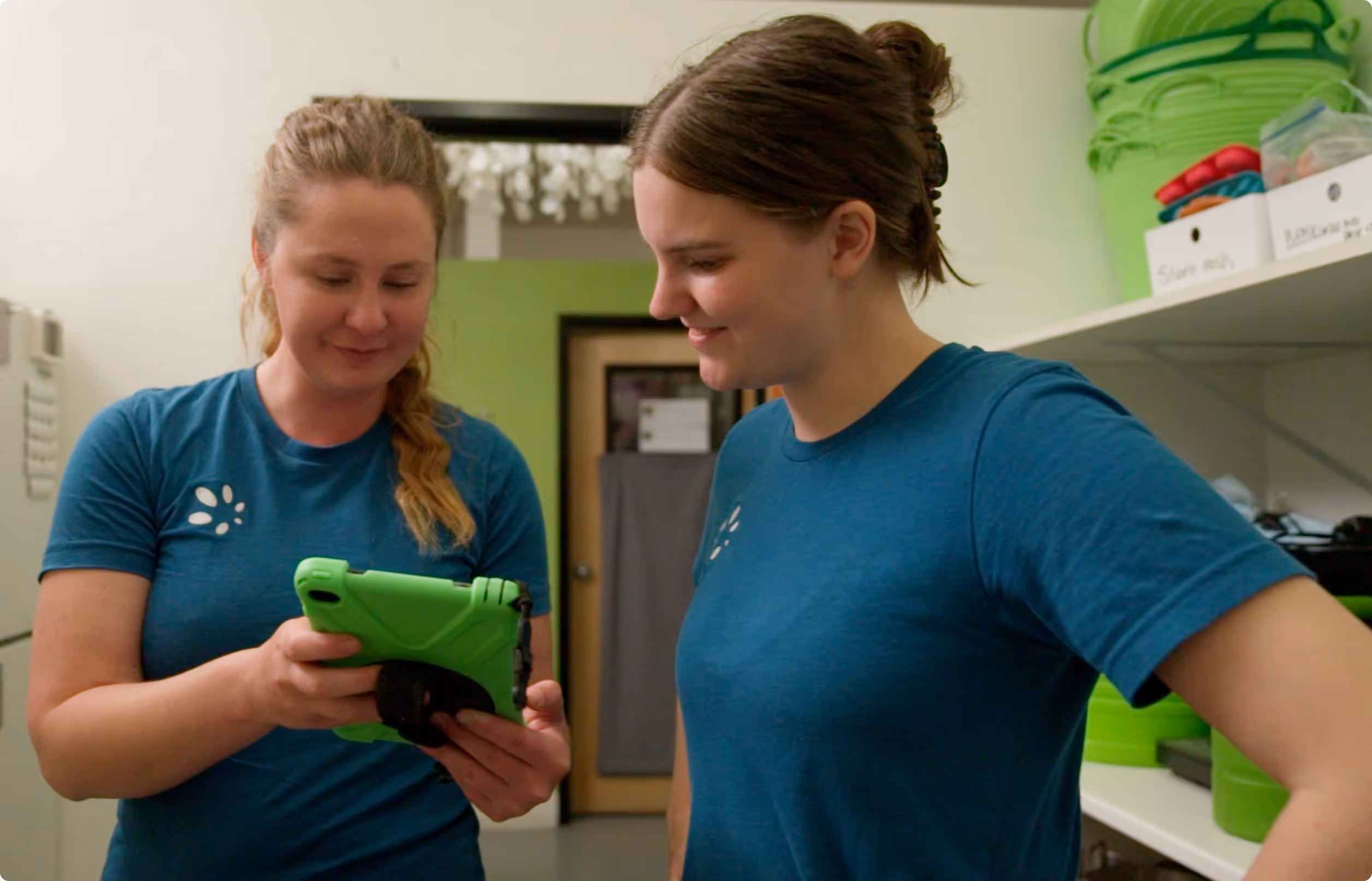 Two women in matching blue shirts looking at a green tablet in a room with shelves and storage bins.