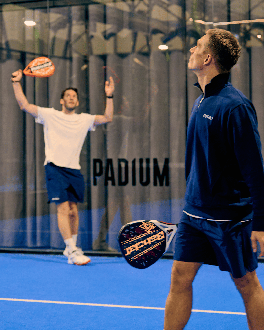 A player holds his racket up to play an overhead shot while a coach in navy watches from the foreground at a Padium court.
